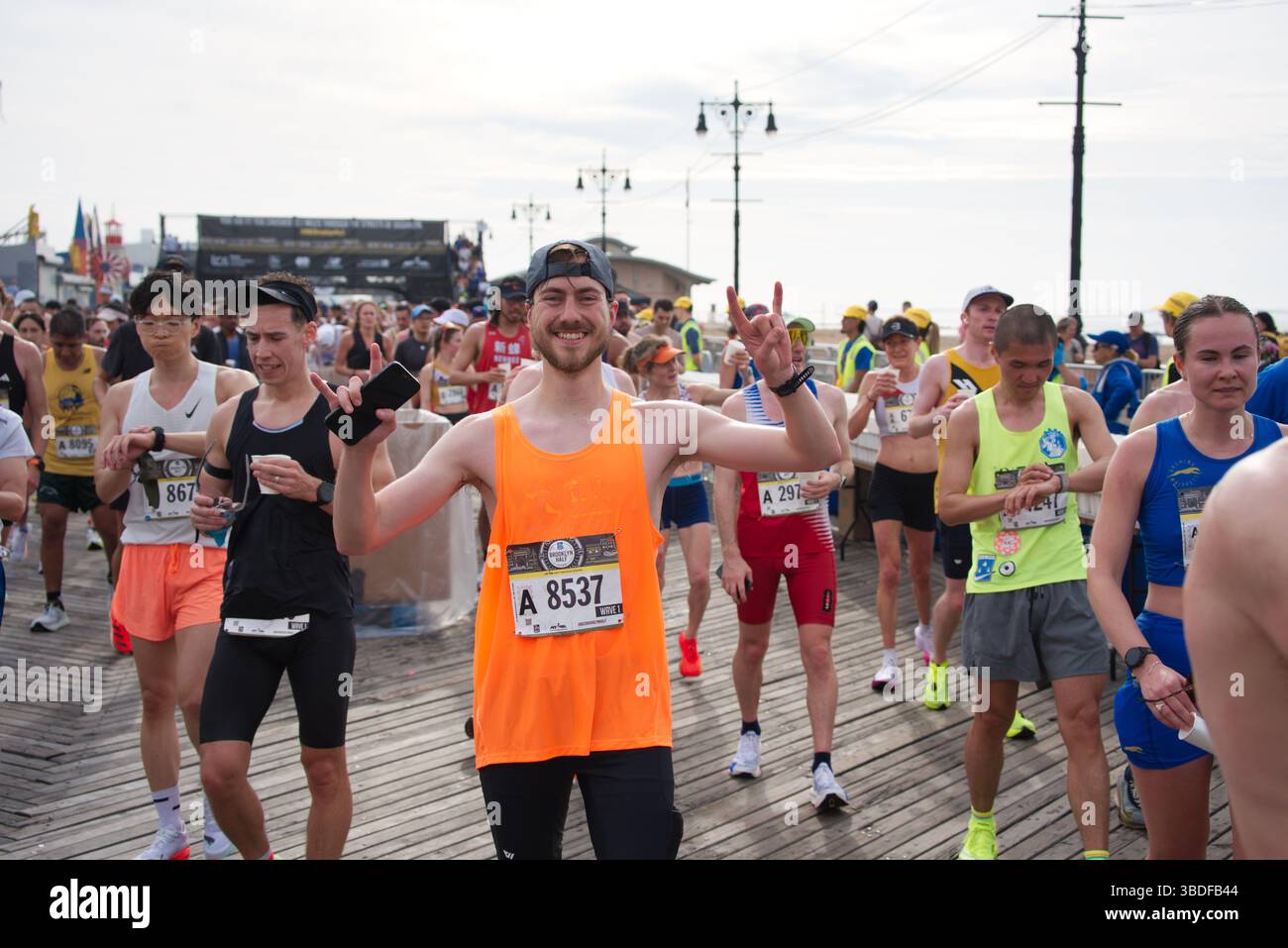Ein lächelnder männlicher Marathonläufer in einem orangen Tanktop, Lätzchen A 8537, mit beiden Händen, umgeben von anderen Läufern auf einer Promenade. Stockfoto