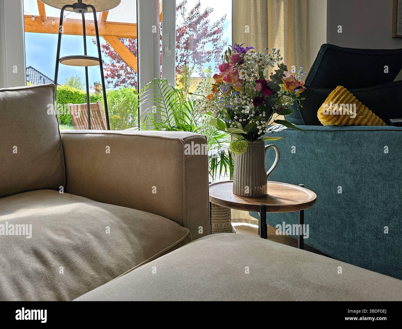 Das sonnendurchflutete Wohnzimmer verfügt über weiche Beige- und Blausitze, einen kleinen Tisch mit einem lebhaften Blumenstrauß und Blick auf den grünen Garten durch Glastüren Stockfoto