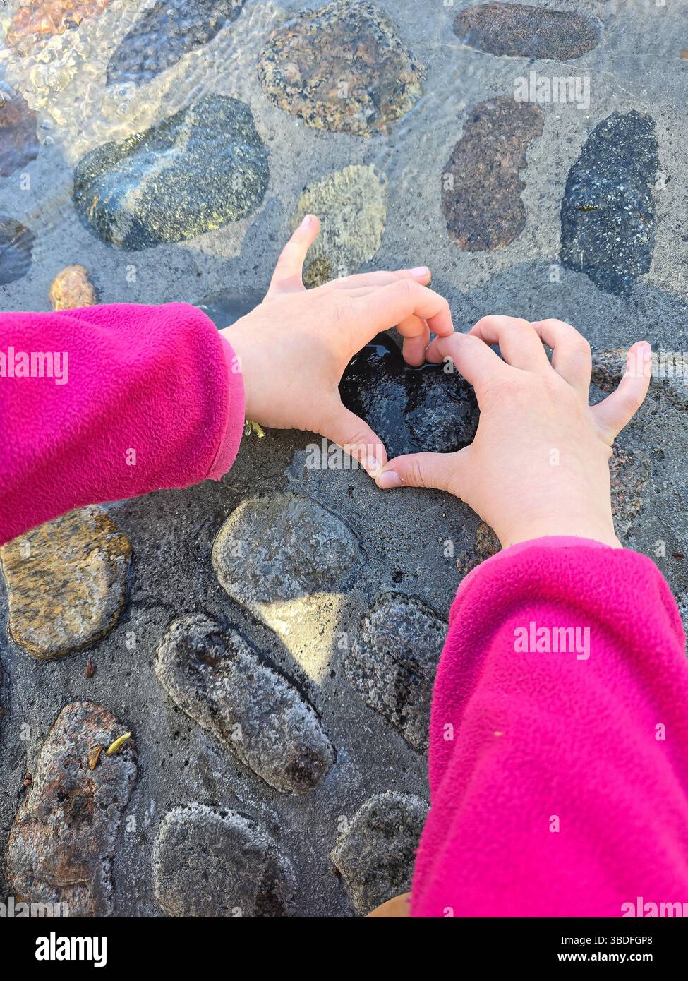 Das Kind in einer rosafarbenen Jacke bildet eine Herzform mit den Händen über einem sonnendurchfluteten Kopfsteinpflasterweg, der Liebe, Sorgfalt und Unschuld symbolisiert Stockfoto