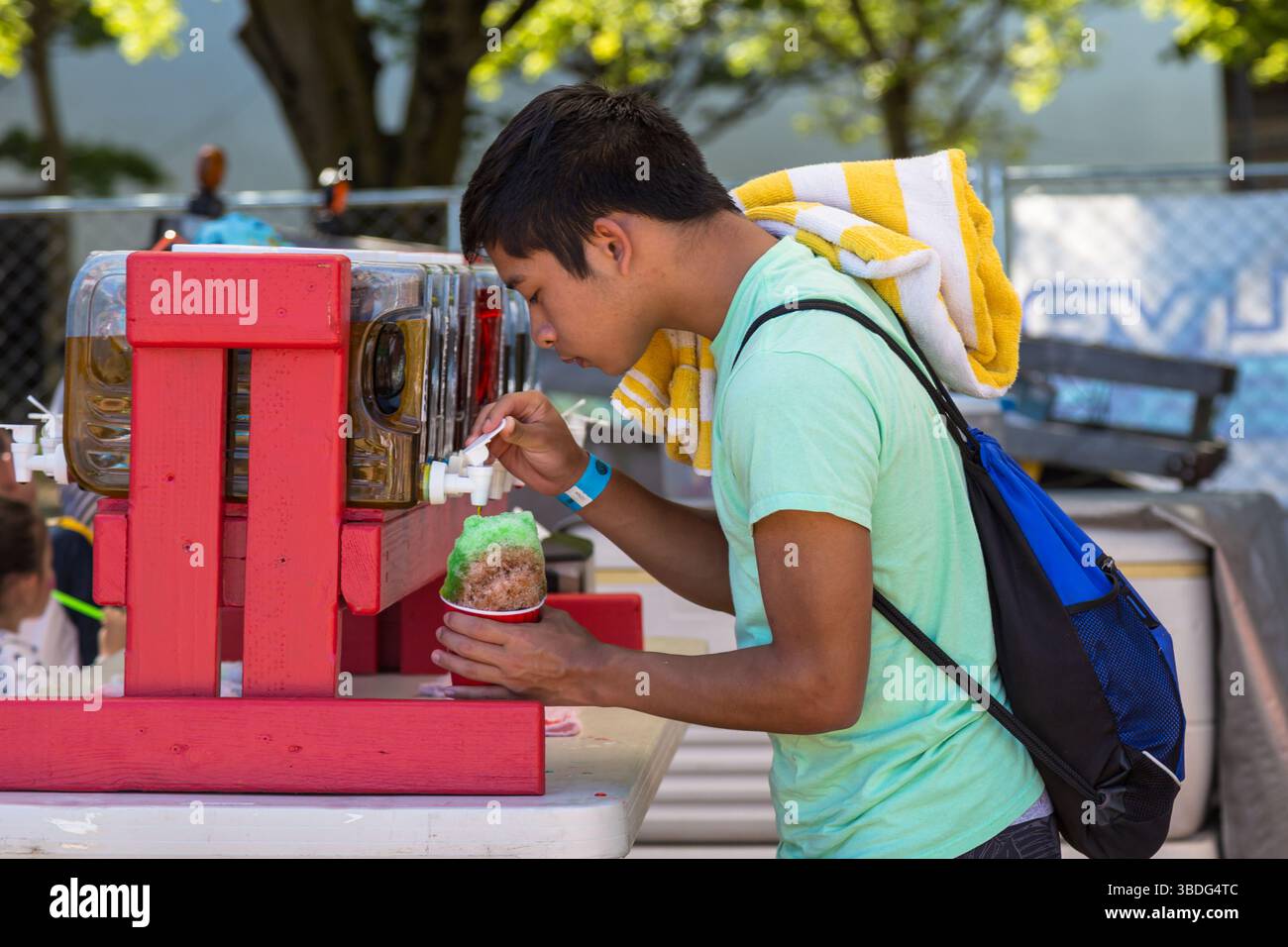 The Big Float, Portland, Oregon - 14. Juli 2018: Ein junger Mann in einem grünen Hemd bekommt einen Schneekegel von einem roten Stand, mit einem gelben Handtuch und einer blauen Tasche. Stockfoto The Big Float, Portland, Oregon - 14. Juli 2018: Ein junger Mann in einem grünen Hemd bekommt einen Schneekegel von einem roten Stand, mit einem gelben Handtuch und einer blauen Tasche. Stockfoto
