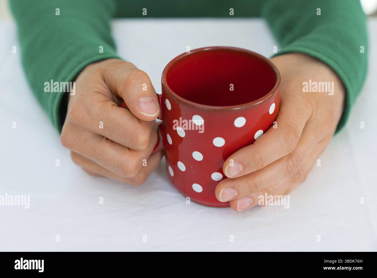 Zarte weibliche Hände einer Frau in einem hellen grünen Pullover holding Eine leere Kaffeetasse Rot mit weißen Tupfen auf einer weißen Tischdecke Stockfoto