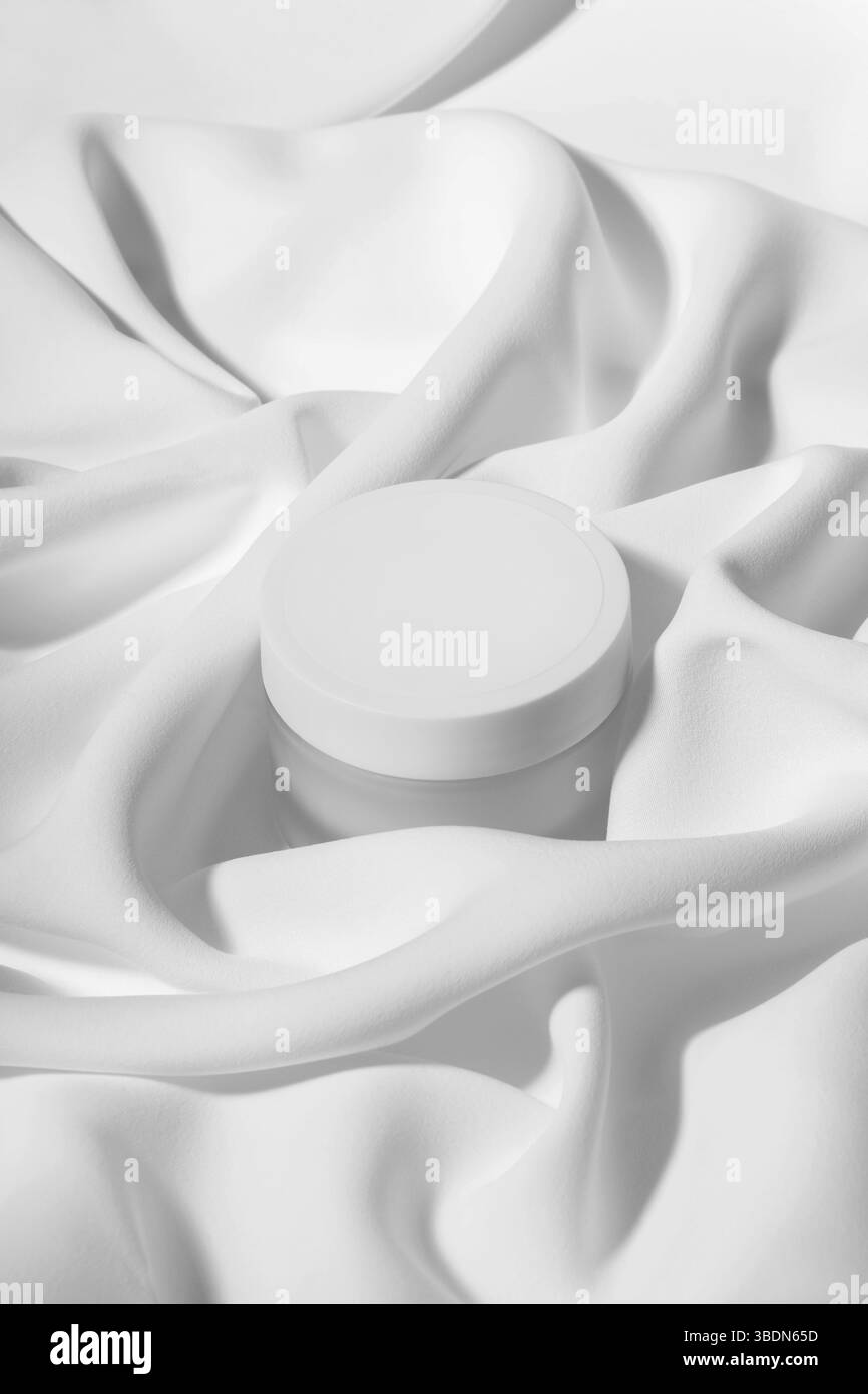 Mockup für das Creme-Logo. Werbefoto von der Leichtigkeit der Creme. Ein rundes weißes Glas Creme zwischen zartem weißem Stoff. Stockfoto