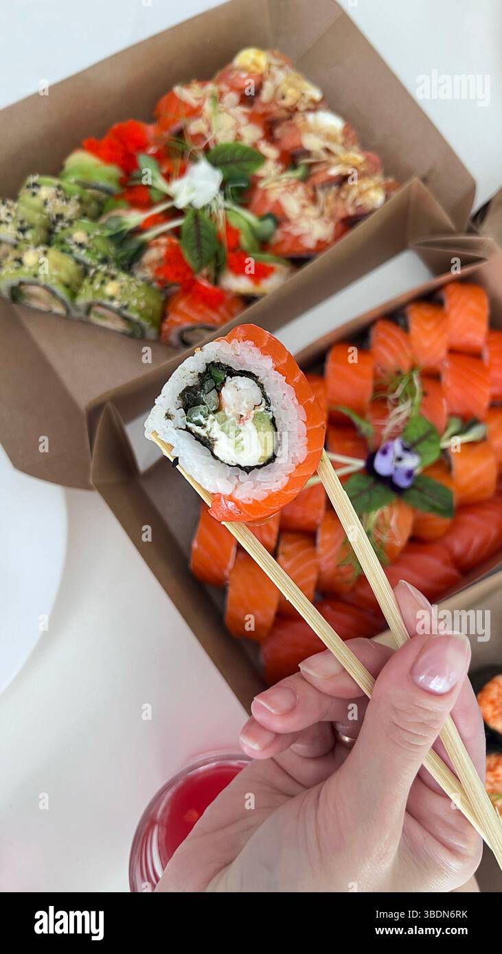 Sushi in der Hand auf dem Hintergrund einer Schachtel mit verschiedenen Sushi. Essstäbchen für Sushi. Sushi-Lieferung. Lifestyle. - Smartphone-aufgenommenes Stockfoto