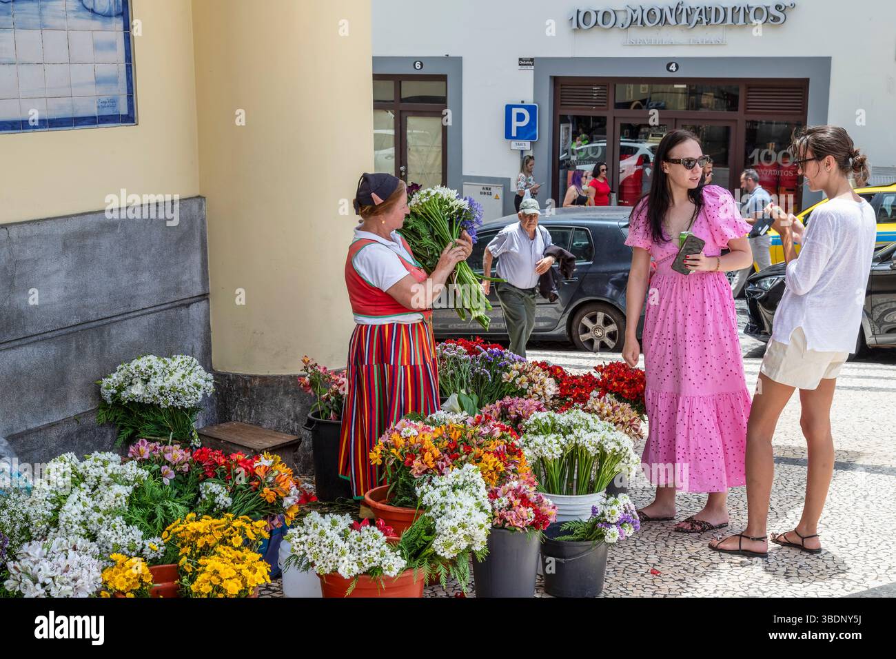 Blumenfrau in rotem Wollrock und weißer Bluse verkauft einen Blumenstrauß an der Straßenecke auf dem Bauernmarkt in Funchal. Stockfoto