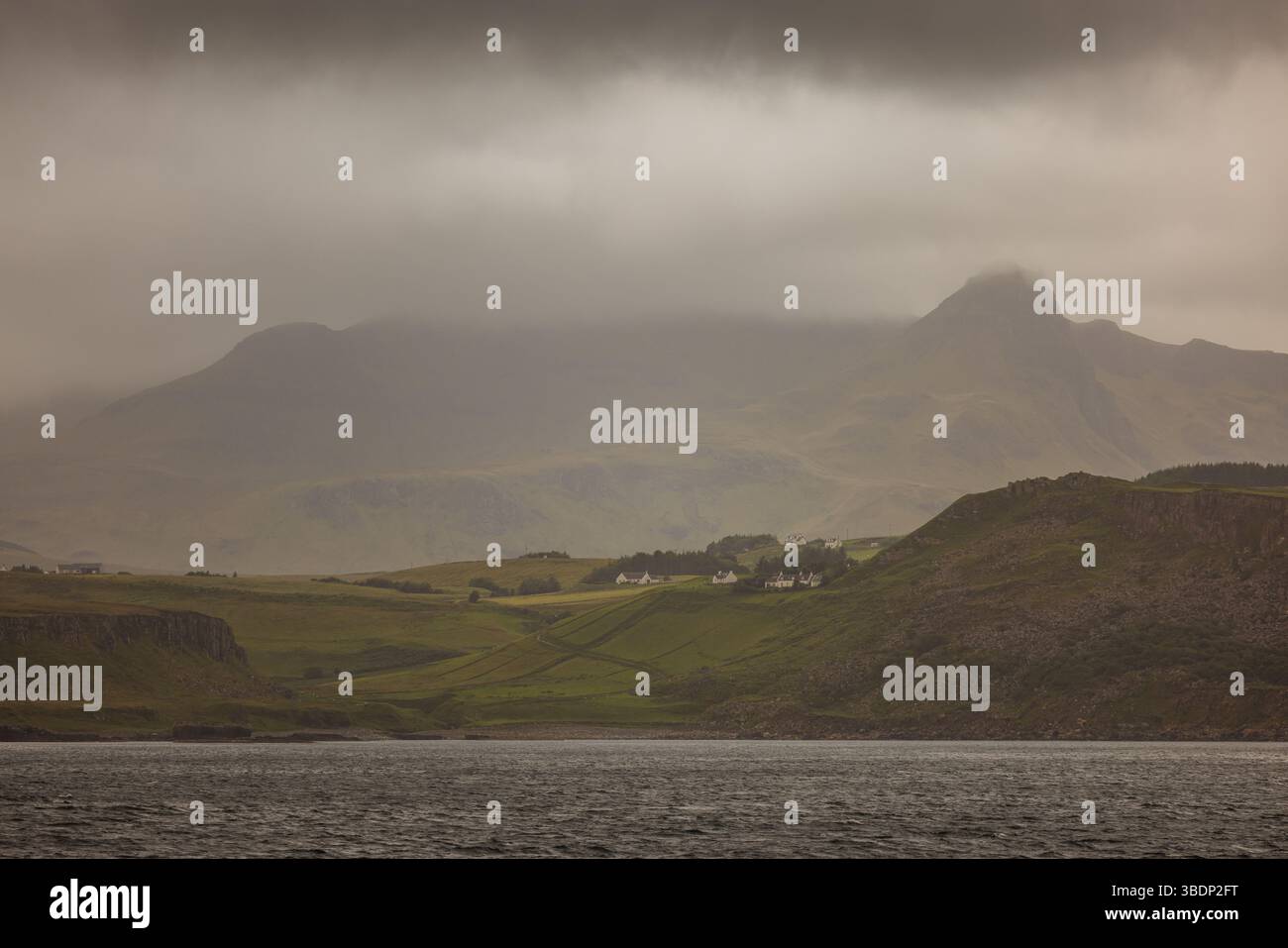 Ländliche Cottages säumen die sanften Hügel unter einem nebelbehüllten Berg auf der Isle of Skye, Schottland, bei weichem Licht und bewölktem Wetter. Stockfoto