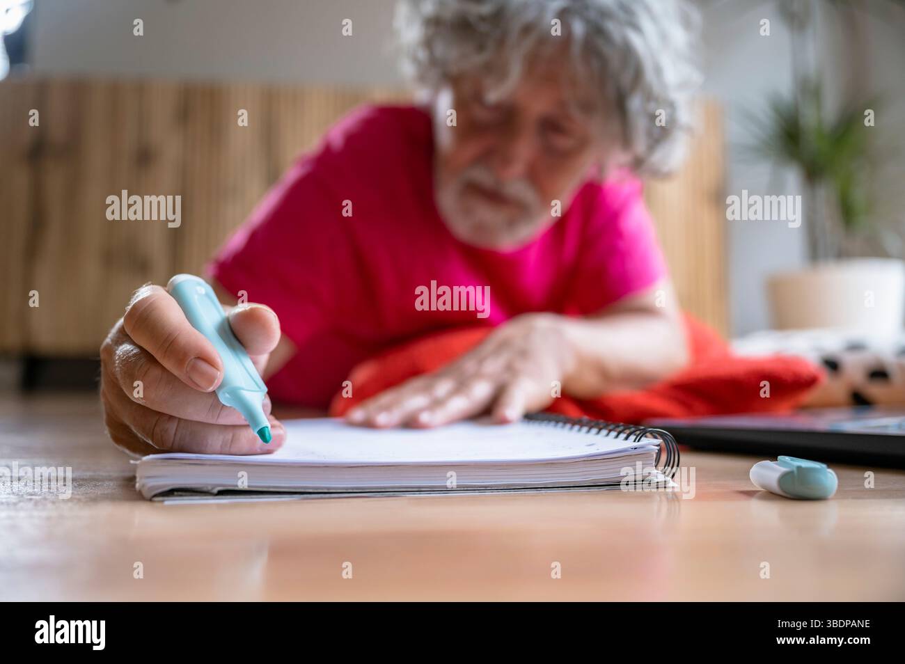Älterer Mann, der auf dem Boden liegt und einen grünen Marker benutzt, um in ein Spiralnotizbuch zu schreiben oder zu zeichnen. Kreative Indoor-Aktivität, Nahaufnahme der Hand, Konzept von Stockfoto