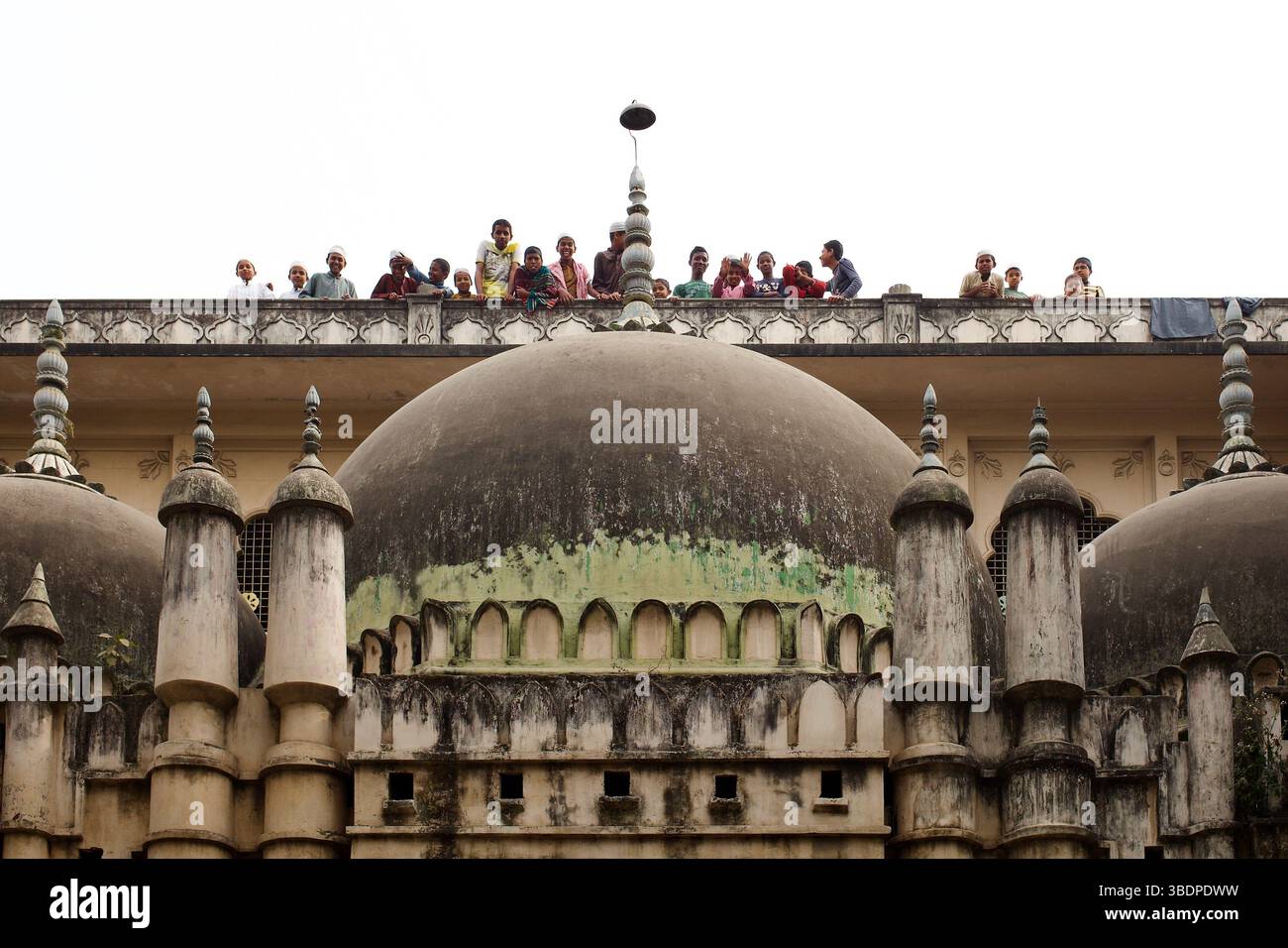 Madrasa-Studenten beobachten von der Dachterrasse eines angrenzenden Gebäudes mit Blick auf die Sat-Gambuj-Moschee aus dem 17. Jahrhundert, Mohammadpur, Dhaka, Bangladesch. Stockfoto