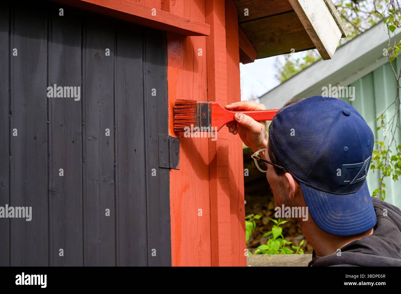 Eine Person trägt eine leuchtend rote Farbe auf ein rustikales Holzgebäude auf, die Handwerkskunst und Liebe zum Detail zeigt. Der Hintergrund zeigt andere Stru Stockfoto