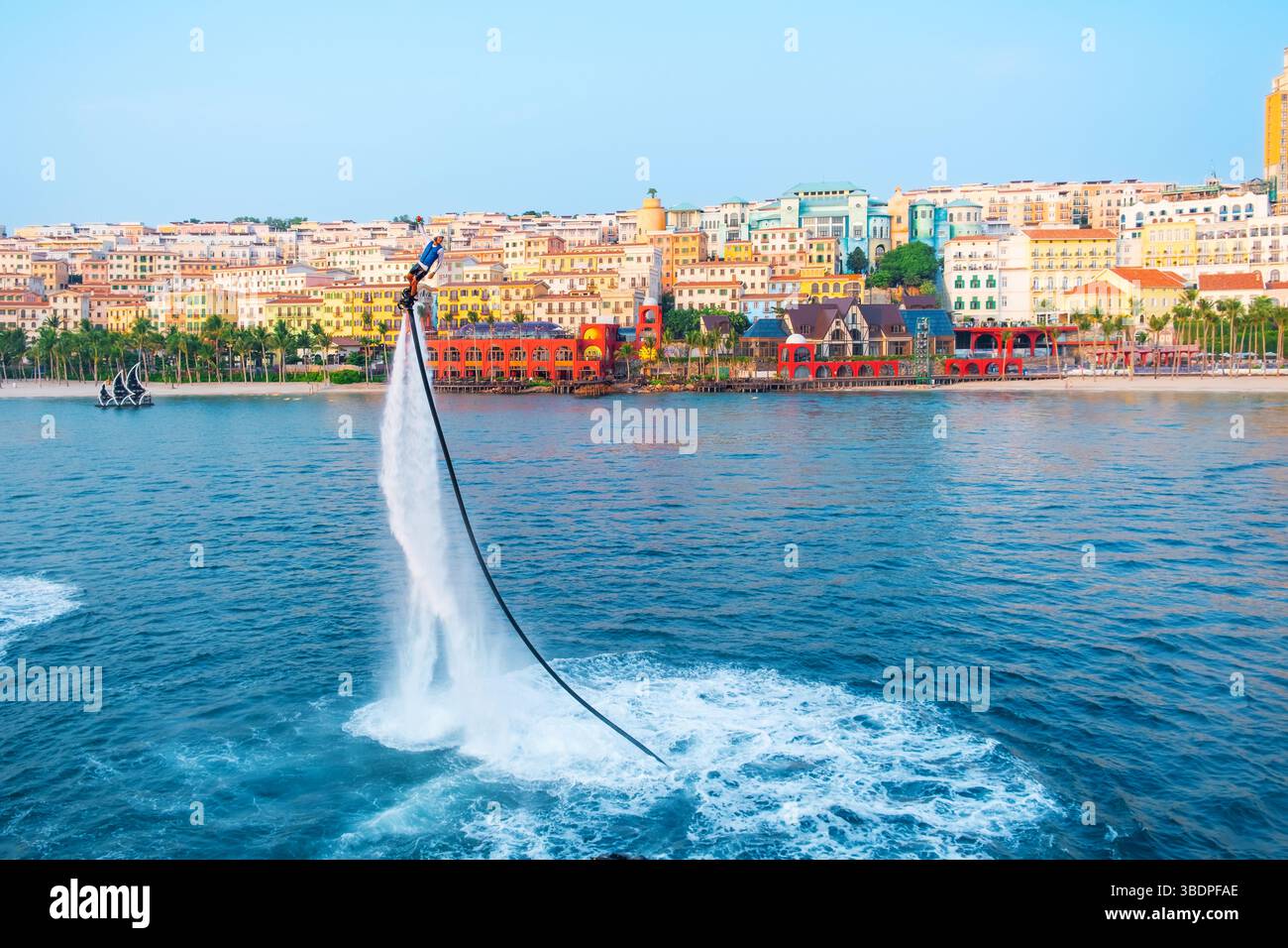 Mann Flyboarding über dem Meer mit farbenfroher Küstenstadt Sonnenuntergang. Extremer Wassersport. Flyboarder ist hoch über dem Wasser und zeigt Action Stockfoto