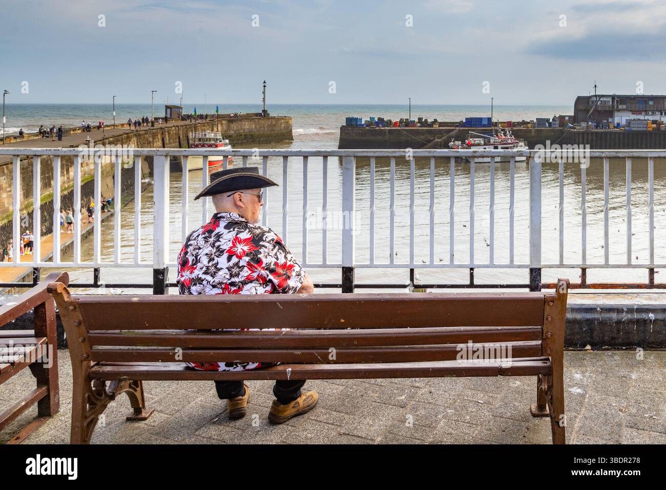 Ein Mann mit Cowboyhut, der auf einer Bank sitzt und über den Hafen von Bridlington blickt. Alleinreisen, Aufenthalt, Urlaub oder Einsamkeit. Stockfoto
