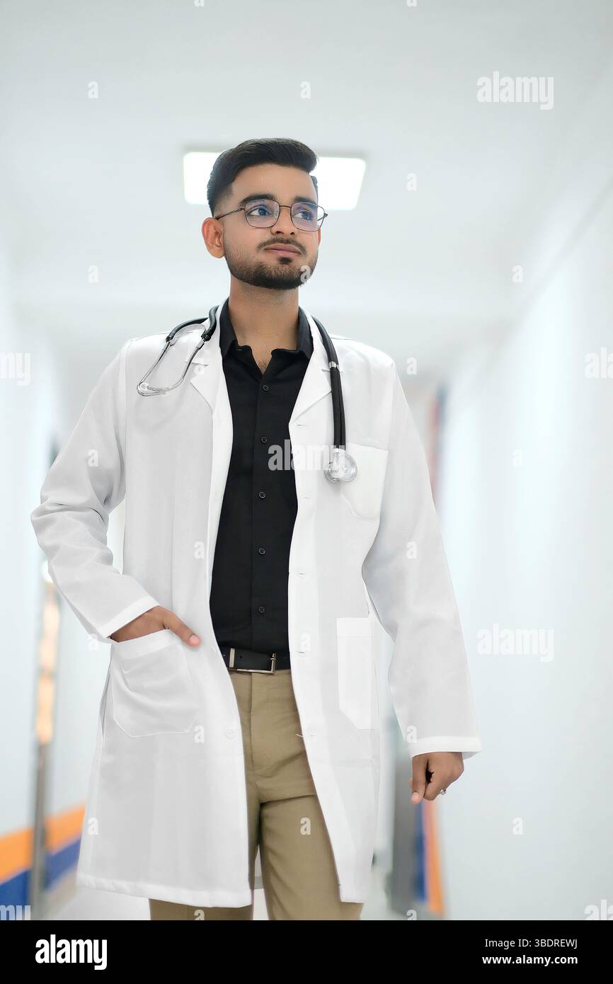 Ein indischer Arzt in einem weißen medizinischen Mantel mit einem Stethoskop um den Hals steht in einem hell beleuchteten Krankenhausflur. Stockfoto