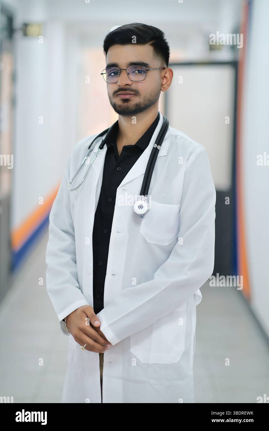Ein indischer Arzt in einem weißen medizinischen Mantel mit einem Stethoskop um den Hals steht in einem hell beleuchteten Krankenhausflur. Stockfoto