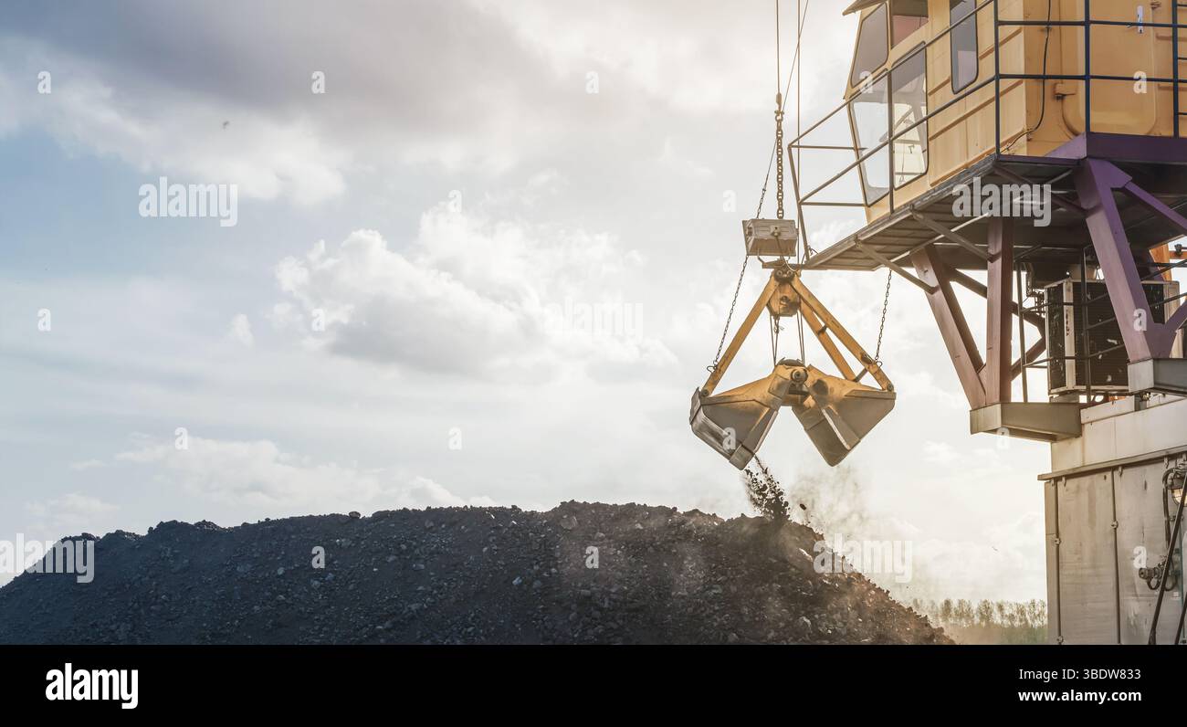 Ein großer gelber Kran schwebt über einem riesigen Kohlehaufen in einem geschäftigen Hafen am Fluss. Wenn die Sonne untergeht, streuen Wolken Licht über die Szene, h Stockfoto