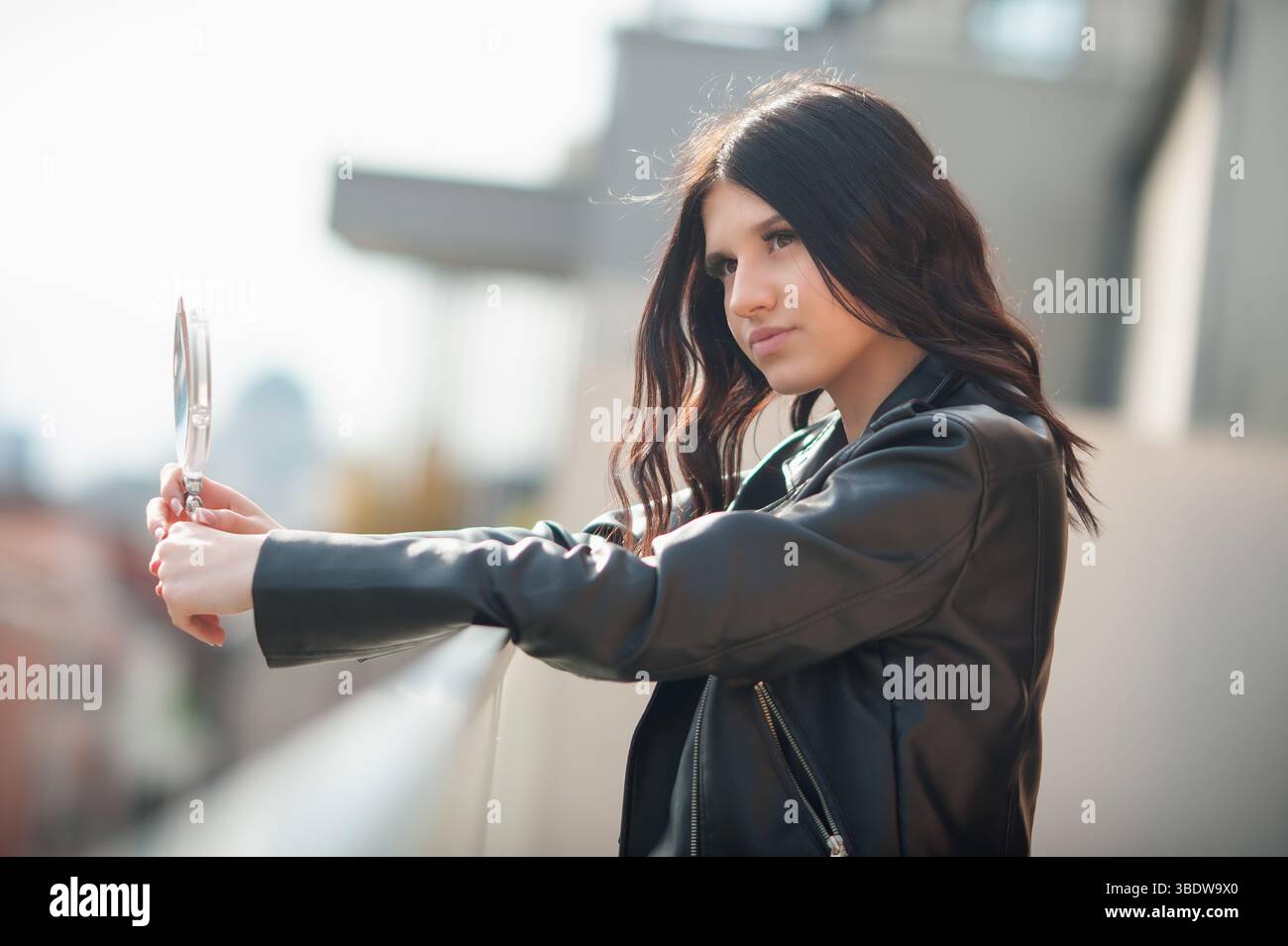 Eine modische junge Frau in einer schwarzen Lederjacke lehnt sich an ein Balkongeländer und blickt in einen Handspiegel. Sie ist auf dem Balkon einer Wohnung Stockfoto