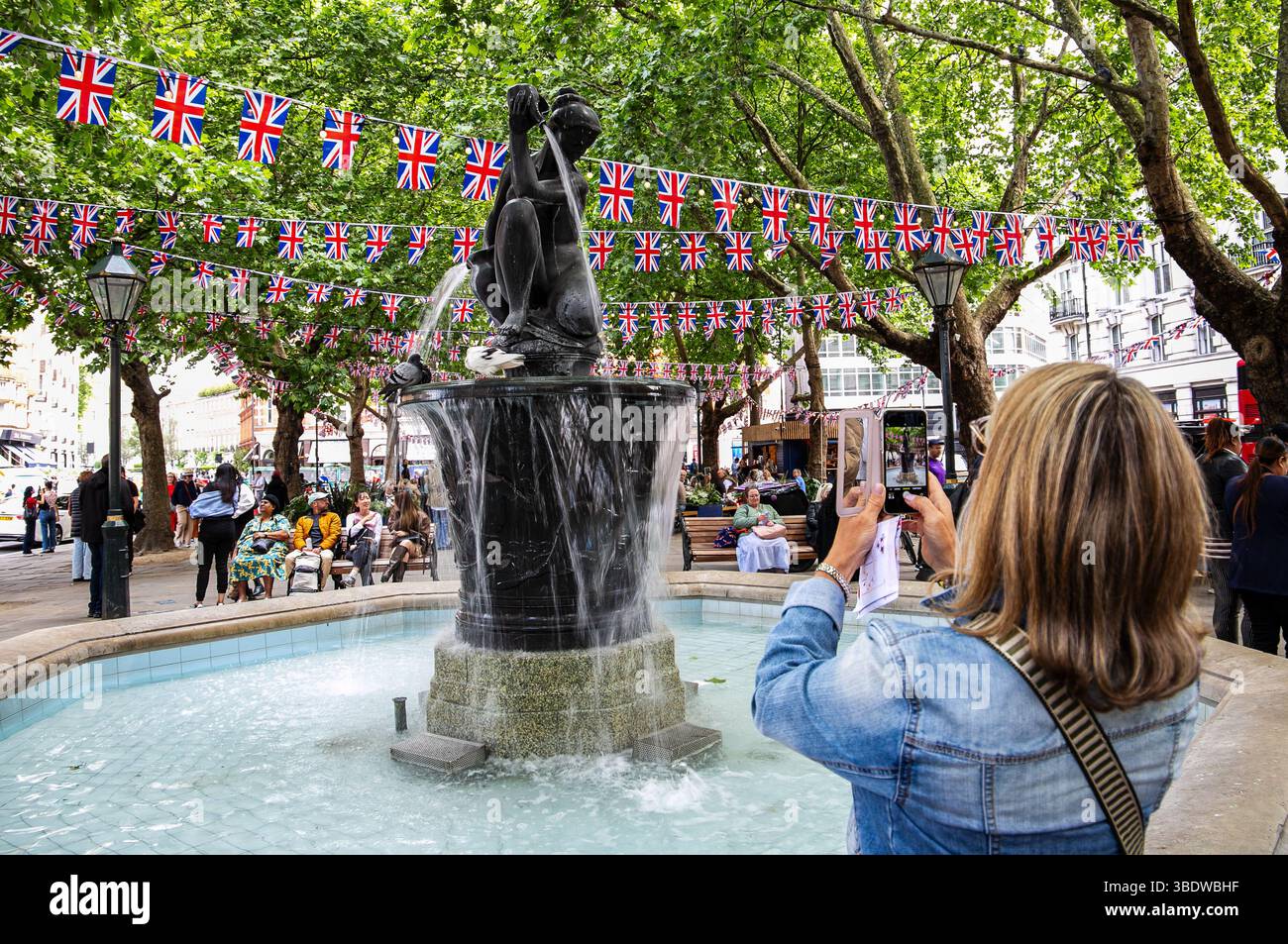 Frau fotografiert einen Brunnen mit einer Statue auf einem belebten Platz, der mit Union Jack Flaggen verziert ist und von People Sloane Square London umgeben ist Stockfoto