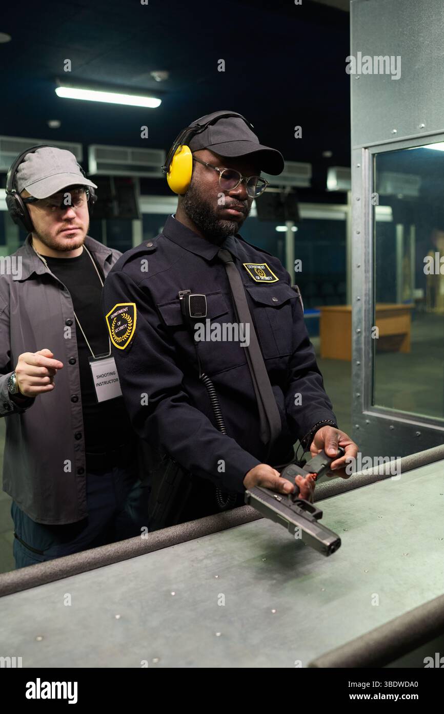 Afroamerikanischer Mann, der auf dem Schießstand im Innenbereich eine Anleitung für die Schusswaffenausbildung erhielt, konzentrierte sich auf den Umgang mit Handfeuerwaffen, wobei der Ausbilder in der Nähe stand und Anweisungen gab Stockfoto