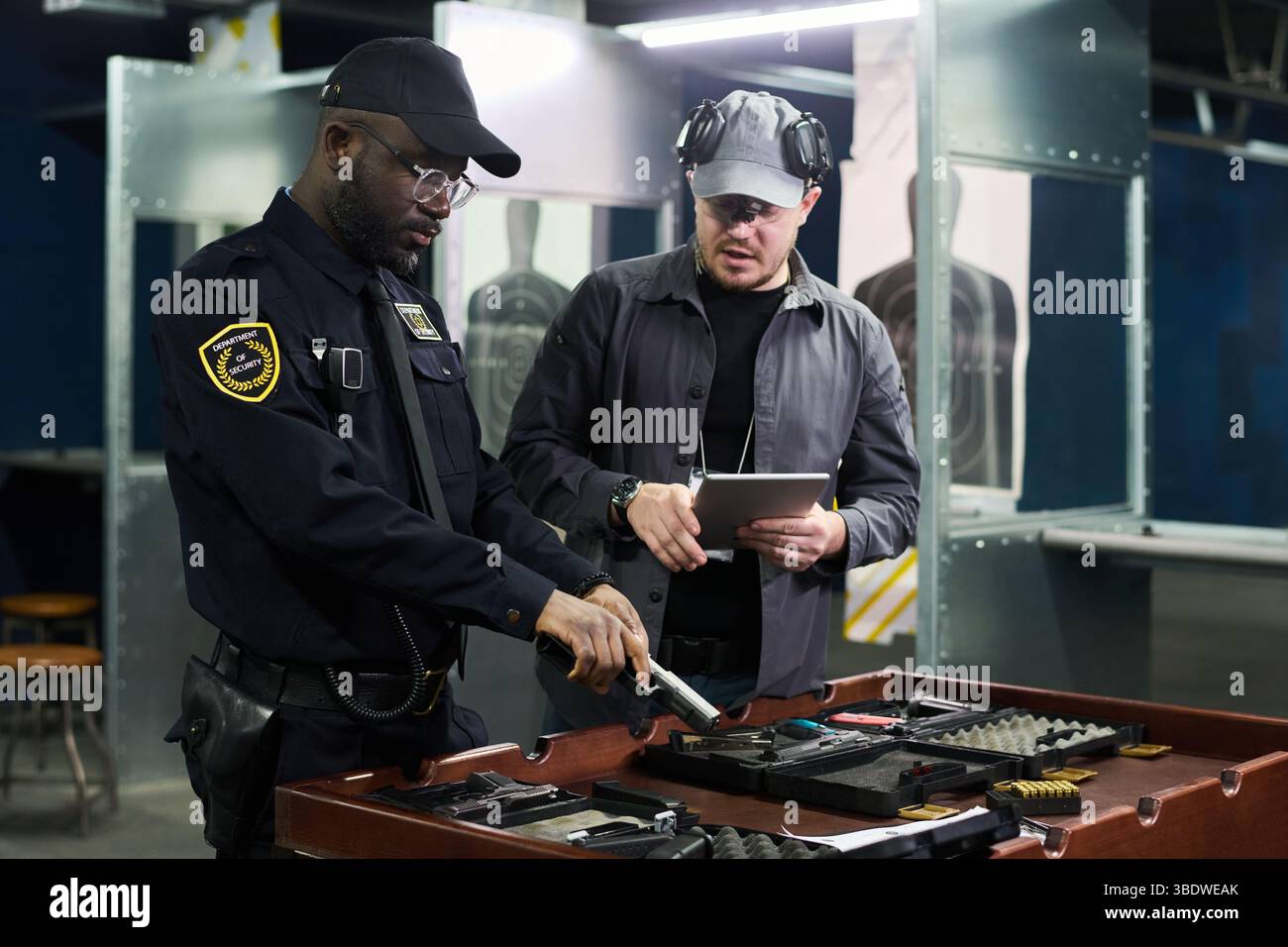 Zwei Männer in Uniform, die auf dem Schießstand zusammenarbeiten, Ausrüstung untersuchen und Notizen auf einem Tablet machen. Hintergrund enthält Zielplatten aus Metall und verschiedene Werkzeuge für die Wartung von Schusswaffen Stockfoto
