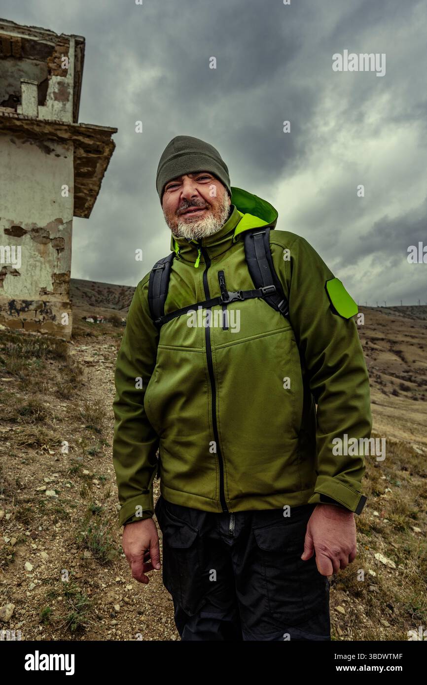 Der Wanderer steht selbstbewusst in der Nähe eines verlassenen Gebäudes vor einem dramatischen bewölkten Himmel in einer bergigen Region Stockfoto