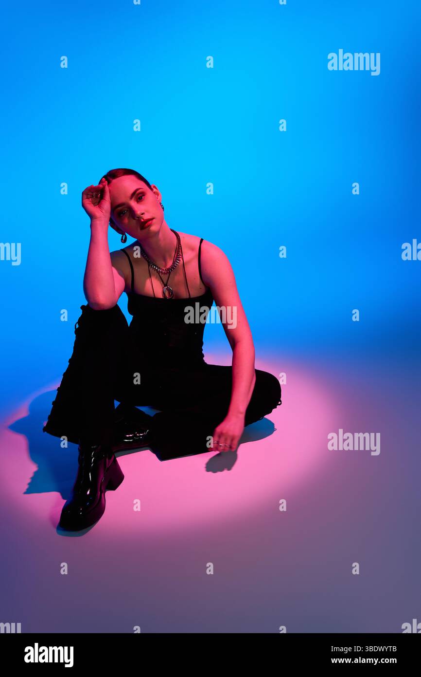 Eine stilvolle junge Frau sitzt anmutig in rosa und blauer Beleuchtung und zeigt moderne Mode. Stockfoto