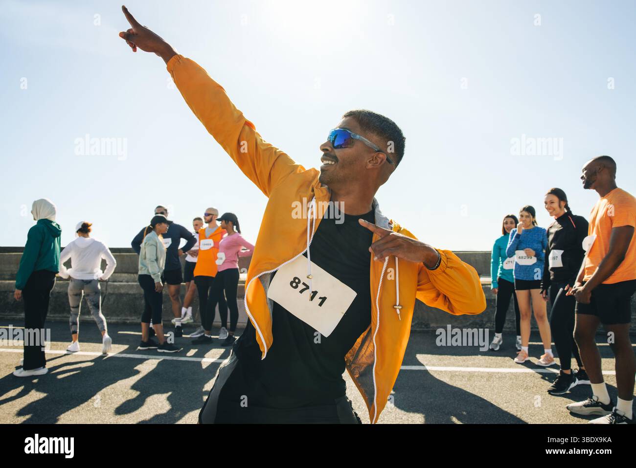 Ein männlicher Athlet mit gelber Jacke posiert bei einem Outdoor-Lauf triumphierend vor anderen Läufern. Die Sonne scheint so hell wie gewohnt Stockfoto