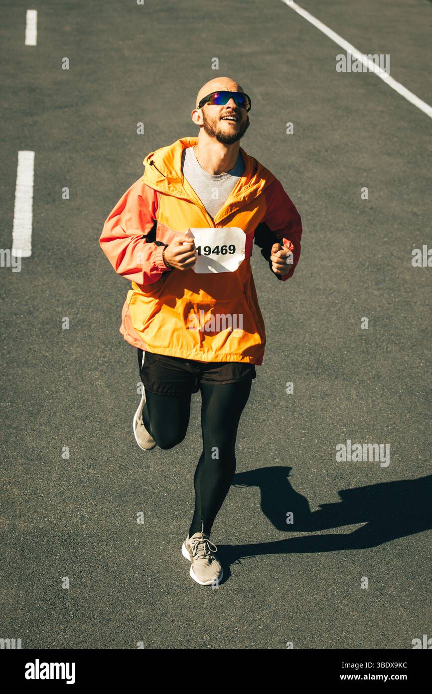 Ein entschlossener Marathonläufer, der eine leuchtend orange Jacke und eine dunkle Laufhose trägt, überwindet auf offener Straße die Ermüdung. Der Ausdruck auf seinem f Stockfoto