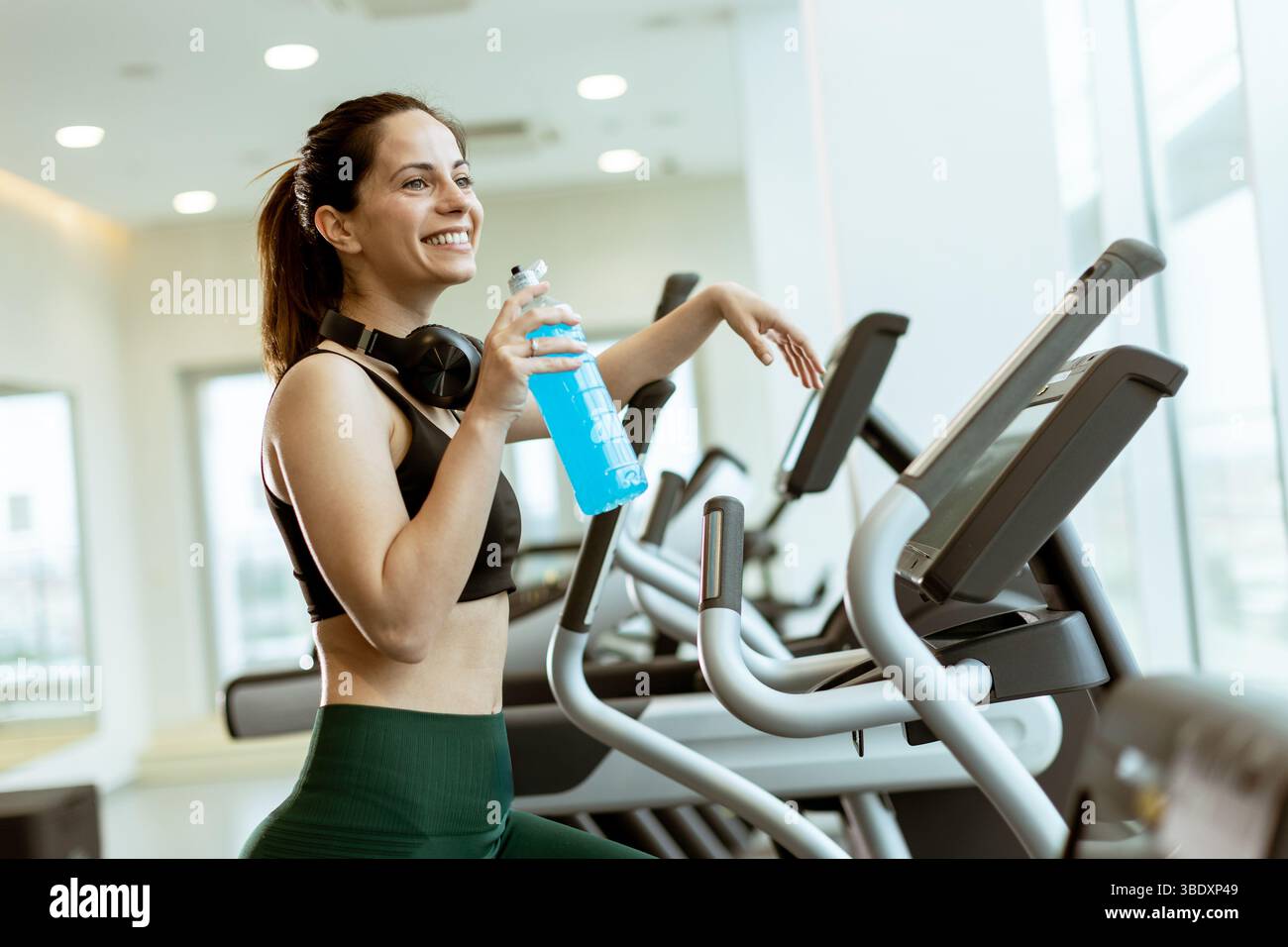 Eine fröhliche Frau hält ihr Training in einem lebhaften Fitnessstudio an, hält ein erfrischendes Getränk und strahlt Energie und Motivation während ihrer Fitnessreise aus. Stockfoto