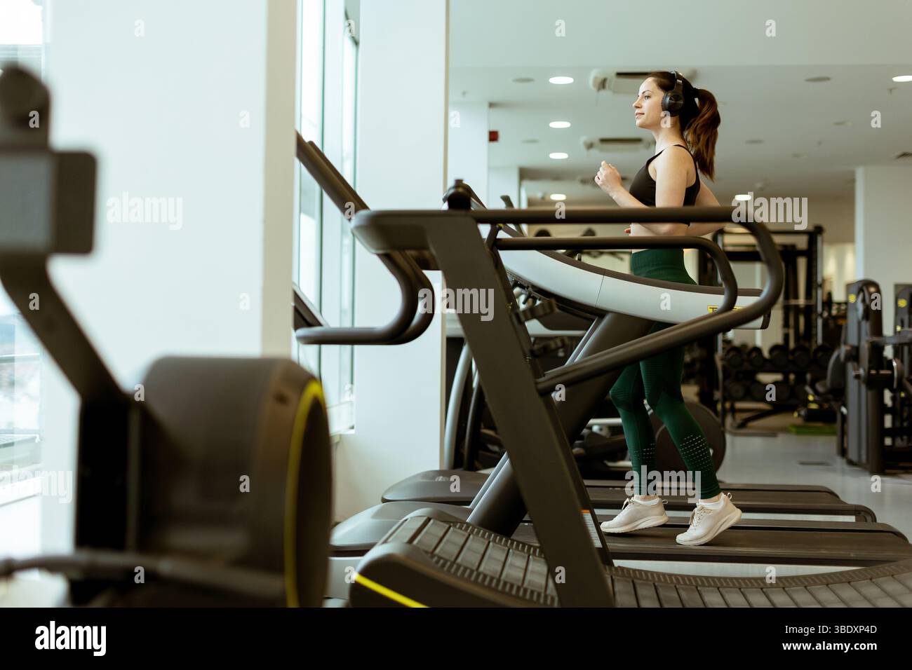Eine konzentrierte Einzelübung auf einem Laufband in einem hellen Fitnessstudio, Kopfhörer an, umgeben von Trainingsgeräten. Stockfoto