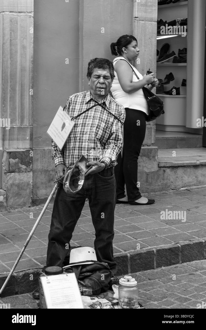 Guanajuato City, Mexiko - 30. Mai 2016: Ein blinder Mann, Miguel, steht auf der Straße mit einem Schild, das um Hilfe bittet, während eine Frau vorbeikommt. Stockfoto