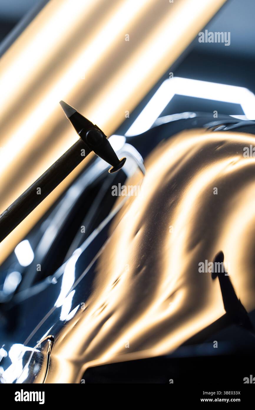 Bei der lacklosen Dellen-Reparatur bei PDR werden Dellen unter Lichtreflexion für Präzision geschlagen. Der Techniker verwendet PDR-Werkzeuge, um Dellen von der Motorhaube zu entfernen Stockfoto