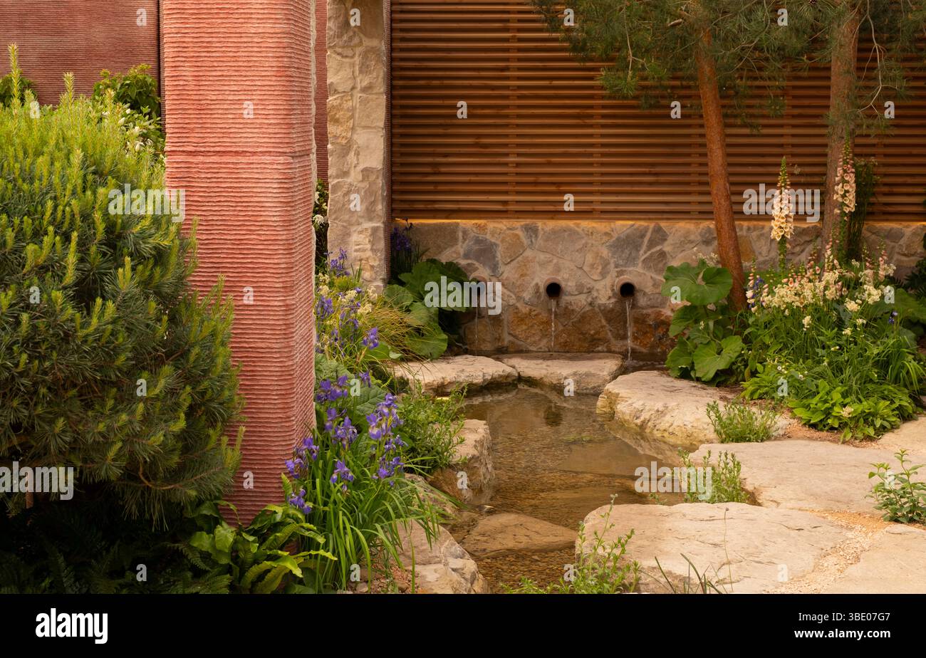 Rohre, die Regenwasser, das in den oberirdischen Trögen gesammelt wurde, in einen Pool leiten, der von Felsen und Blumen umgeben ist, im Killik & Co 'Save for a reiny Day' Garden d Stockfoto