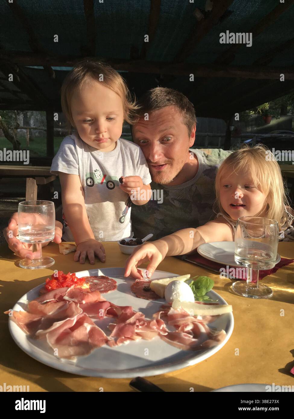Glückliche Familie genießt einen Teller italienische Antipasti während eines Picknicks im Freien in der Sonne. Zwei Söhne und ein Vater schauen mit Begeisterung auf die Vielfalt von Stockfoto