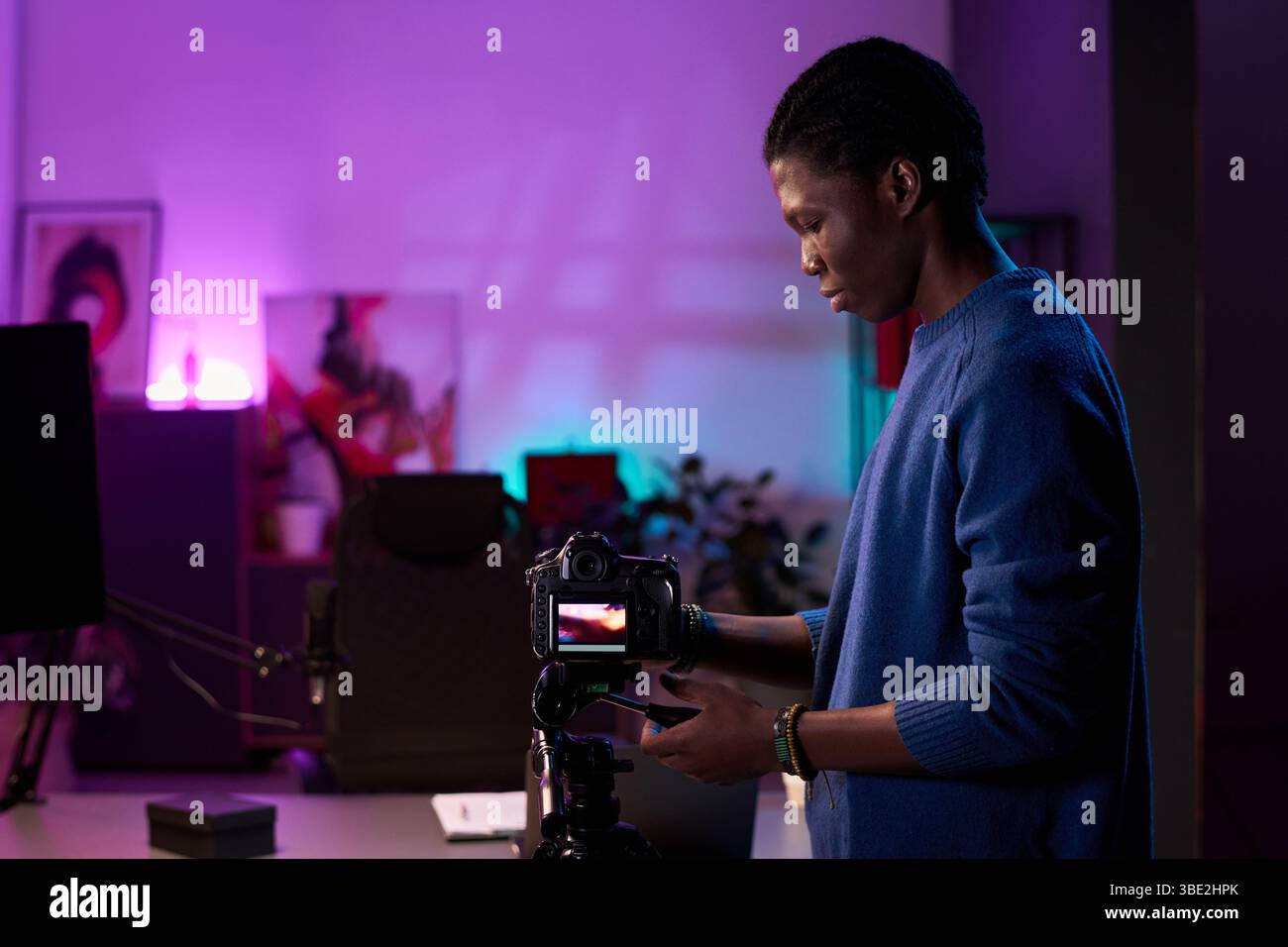 Fotograf im Studio passt die Kameraausrüstung auf einem Stativ mit farbenfrohen Hintergrundlichtern und Bürostuhl im Rahmen an und schafft so eine dynamische Arbeitsumgebung Stockfoto