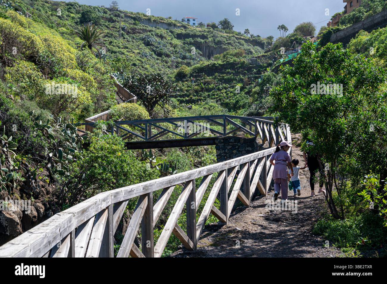 Eine Frau und ein Kind gehen auf einer Holzbrücke über einen Weg. Die Brücke ist von Bäumen und Büschen umgeben und der Himmel ist bewölkt. Stockfoto