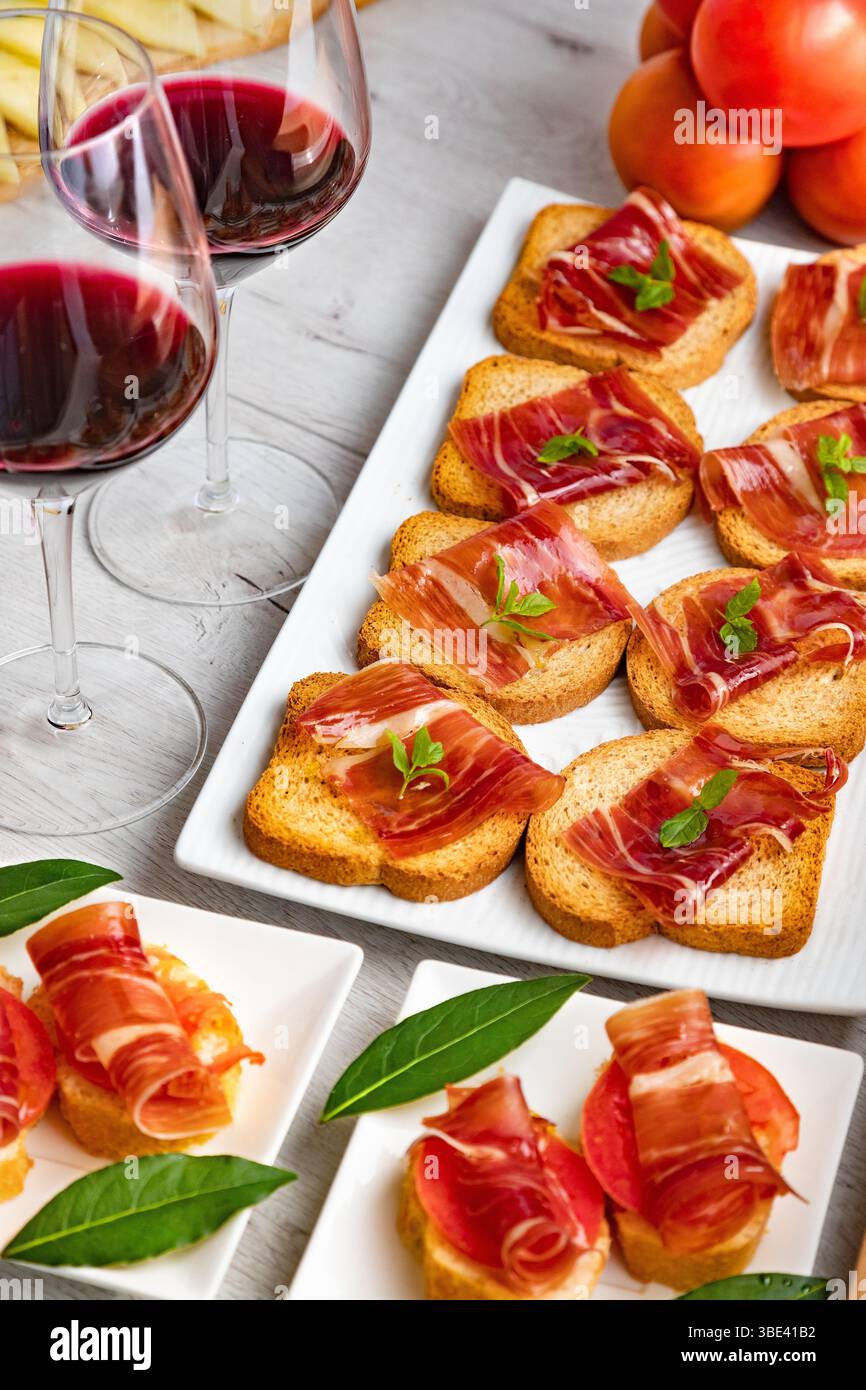 Tapas und Vorspeisen mit iberischem Schinken mit einem Glas Wein, Reifen Tomaten und Kuhkäse auf weißem Holzhintergrund. Spanische Tapas und Vorspeisen Stockfoto