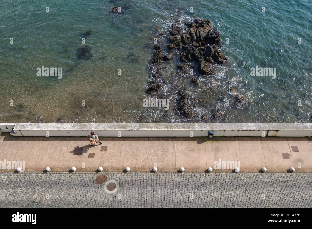 Blick von oben auf die Küstenpromenade mit einer Person, die neben der Rocky Shore läuft. Ozeanwasser, Seawall, Städtische Küste, Freizeitaktivitäten Stockfoto