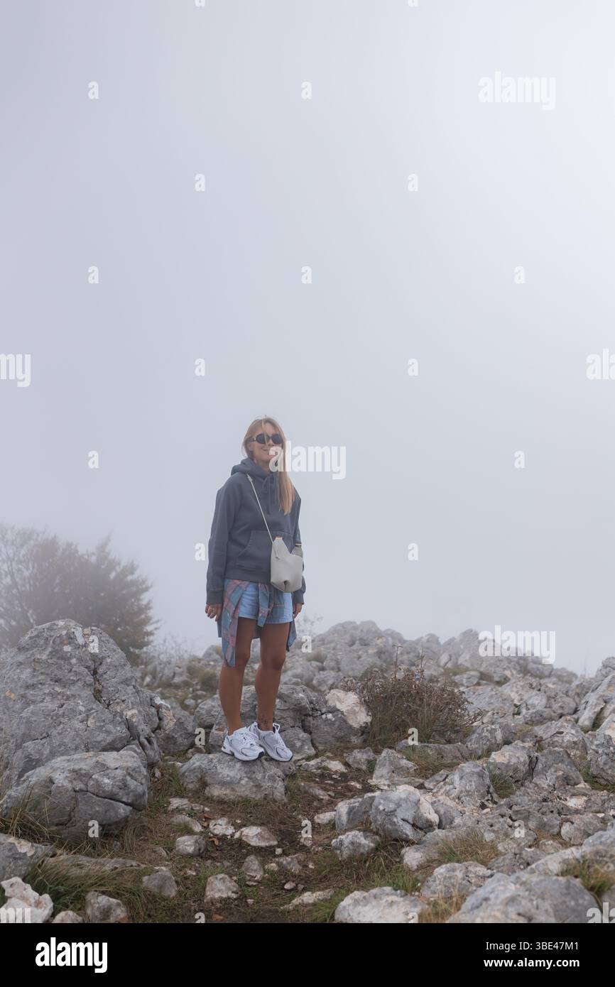 Die Frau blickt in lässigem sportlichem Outfit mit weißem Crossbody leicht nach unten, während sie auf einem nebeligen, felsigen Hügel steht. Stockfoto