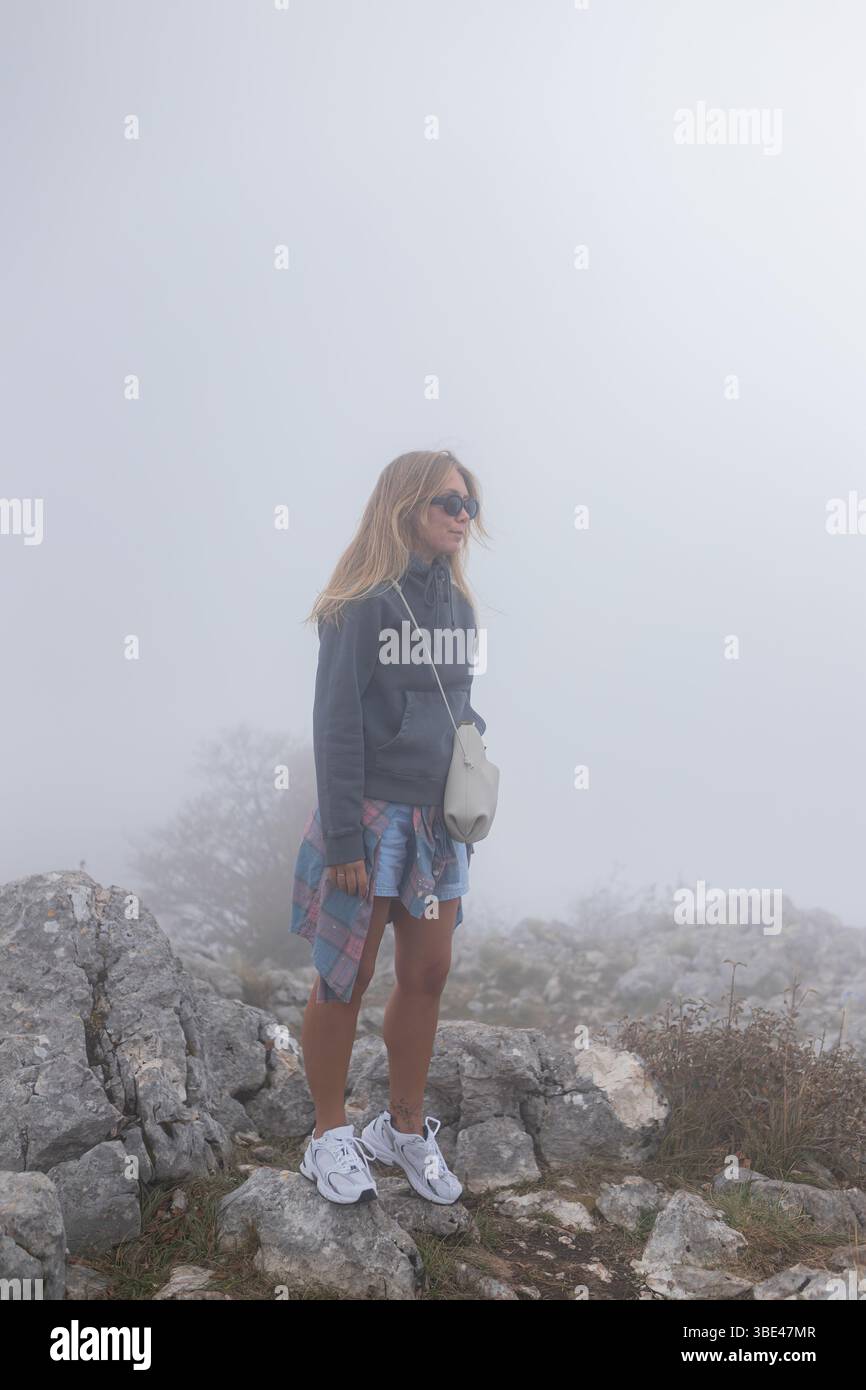 Frau auf einem nebligen felsigen Hügel mit langen blonden Haaren und sportlichem Outfit mit friedlichem Blick auf die Berge. Stockfoto