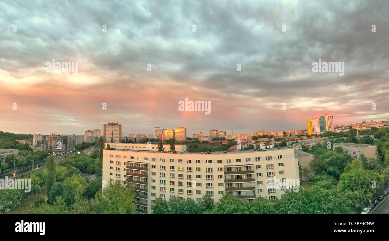 Farbenfrohe Sonnenuntergangswolken über einer üppig grünen städtischen Umgebung mit sanftem Regenbogen. Hohe Gebäude und Wohngebiete schaffen eine Mischung aus Natur und Mode Stockfoto