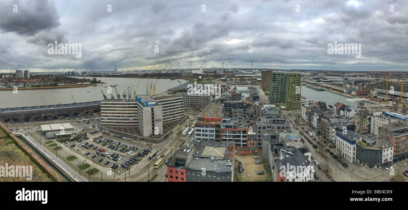 Das Weitwinkelfoto erfasst ein lebhaftes Stadtbild am Fluss mit modernen Gebäuden und industriellen Elementen unter einem dramatischen bewölkten Himmel. Stockfoto