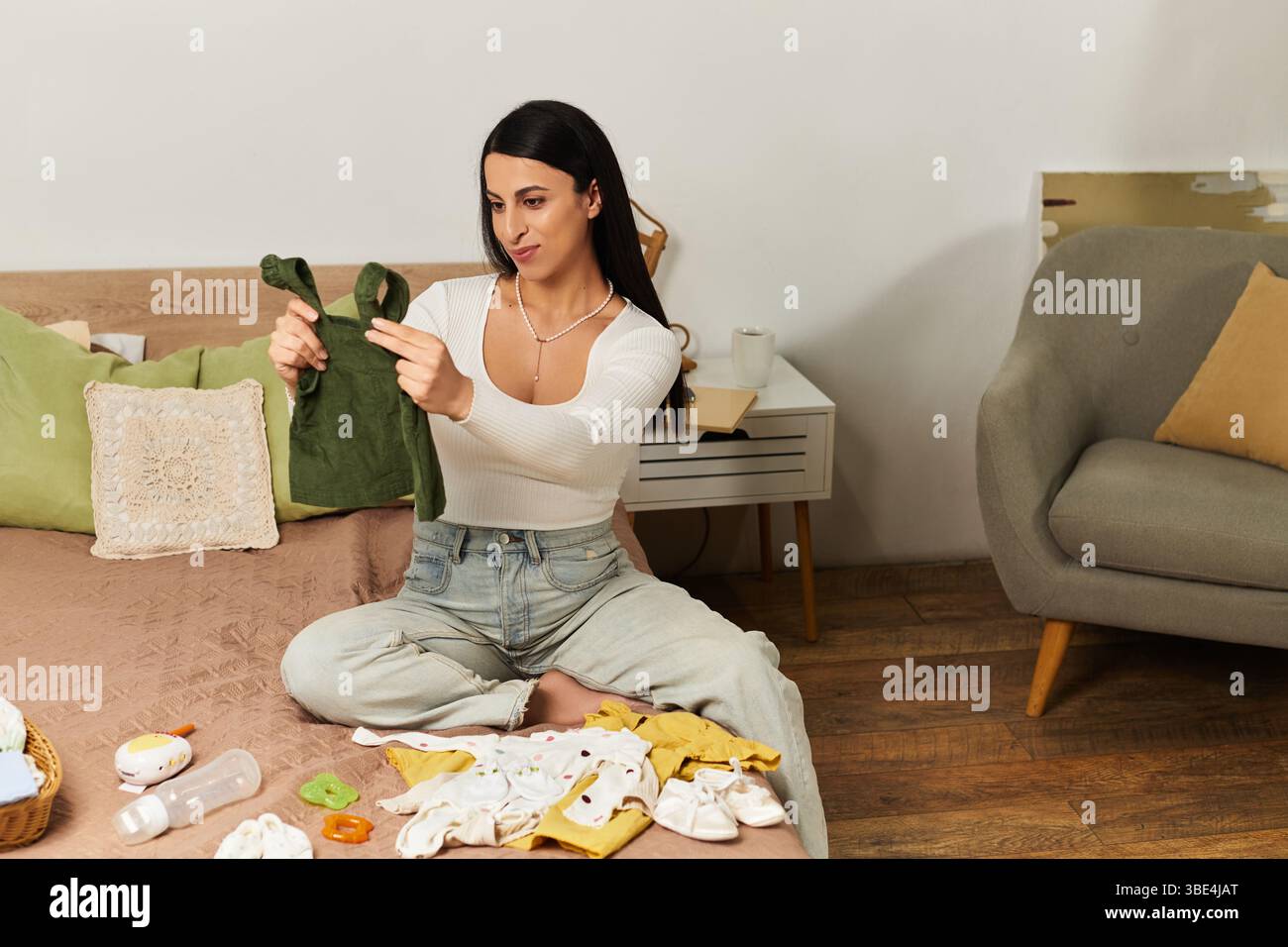 Eine schwangere Frau sitzt auf einem Bett und sortiert Babykleidung in einem friedlichen Moment. Stockfoto