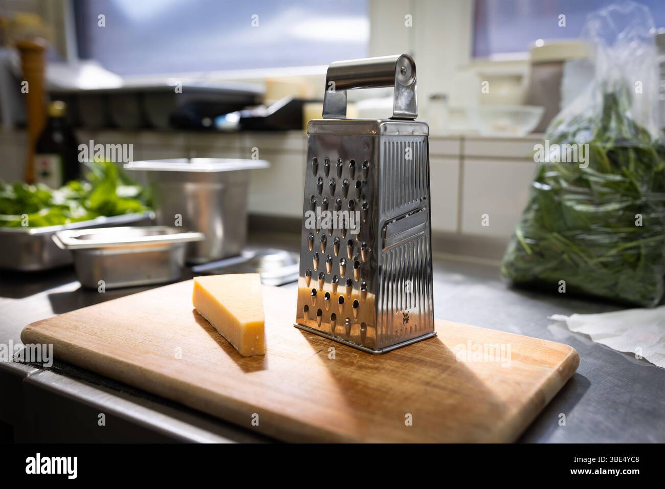 Deutschland 03. April 2025: Im Bild: Eine robuste Küchenreibe aus glänzendem Edelstahl steht im Fokus auf einem hellen Holzschneidebrett, bereit für die Zubereitung von Speisen. Daneben liegt ein appetitliches Stück Hartkäse, der nach Parmesan oder Grana Padano aussieht und auf seinen Einsatz warten. Diese Szene spielt sich in einer professionellen Küchenumgebung ab, erkennbar an den Edelstahlflächen, den Fliesenwänden und weiteren Küchenutensilien wie eine Flasche Olivenöl und einem Beutel frischer grüner Blätter im Hintergrund. Das Bild fängt die Essenz der handwerklichen Lebensmittelzubere Stockfoto