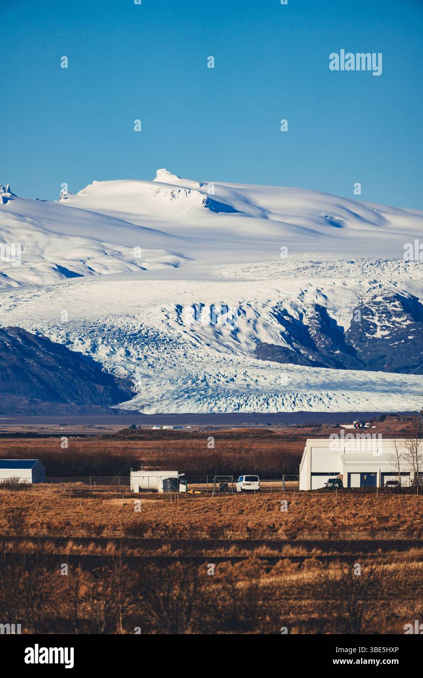 Fernansicht eines Gletschers aus der Gegend von Stokksnes in Island, mit kleinen ländlichen Gebäuden und goldenen Feldern im Vordergrund unter einem hellblauen Himmel. Stockfoto