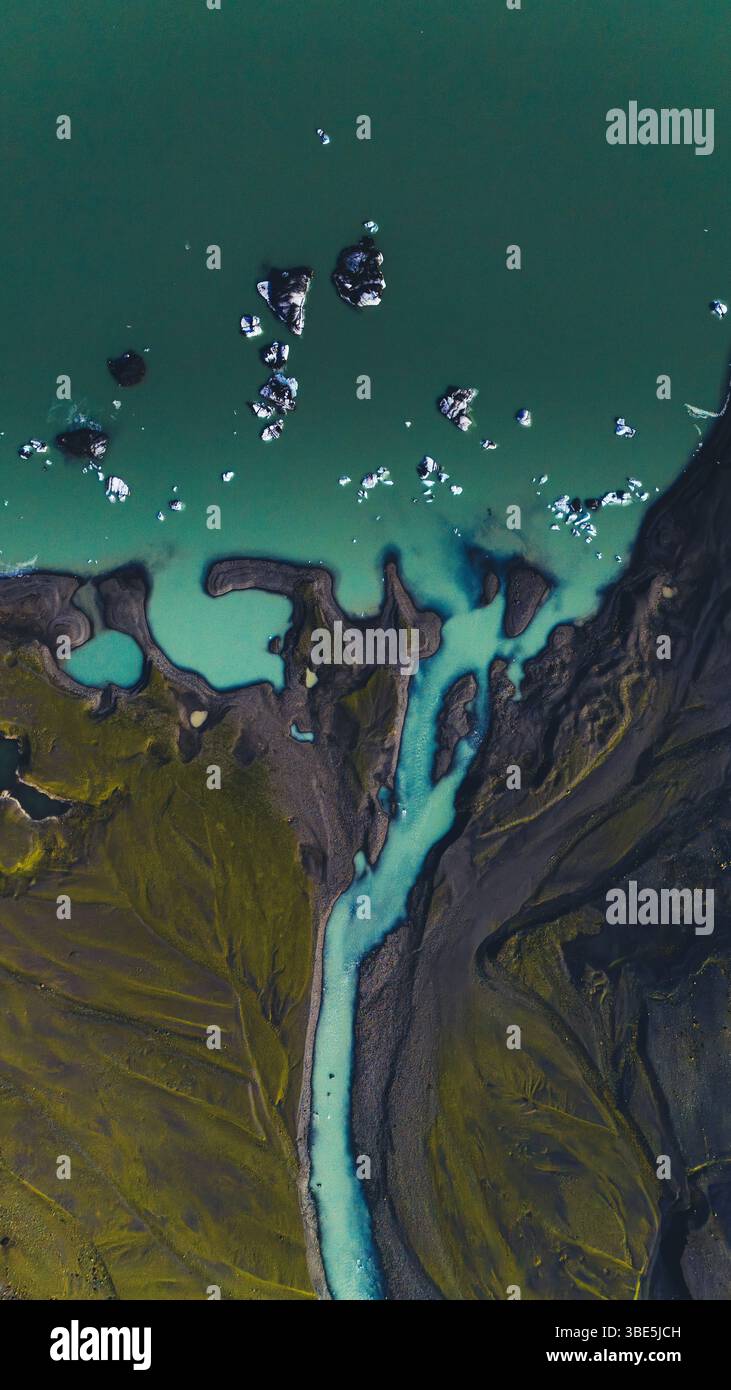 Vertikale Drohnenaufnahme eines geflochtenen Gletscherflusses, der auf eine grüne Lagune trifft, mit Eisbergen, die in der Nähe des Ufers schwimmen. Eine lebendige Luftaufnahme von Islands roher gla Stockfoto