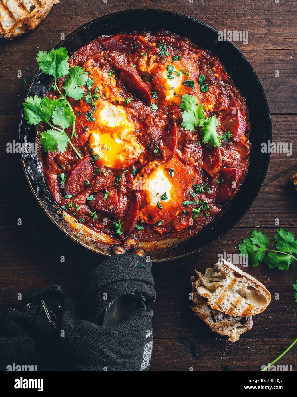 Genießen Sie die lebendigen Farben und das verlockende Aroma eines traditionellen Shakshuka-Gerichts mit pochierten Eiern in würziger Tomatensauce, garniert mit frischen Kräutern und Sesa Stockfoto