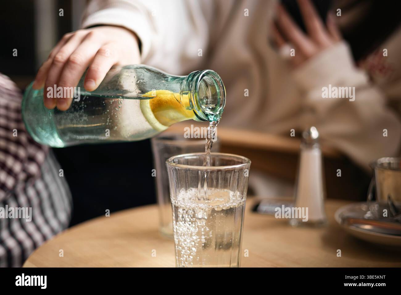 Das zugeschnittene, nicht erkennbare Mädchen gießt Wasser aus einer Flasche mit einem Stück Zitrone in ein Glas in einem Café und fängt einen Moment der Erfrischung ein. Stockfoto