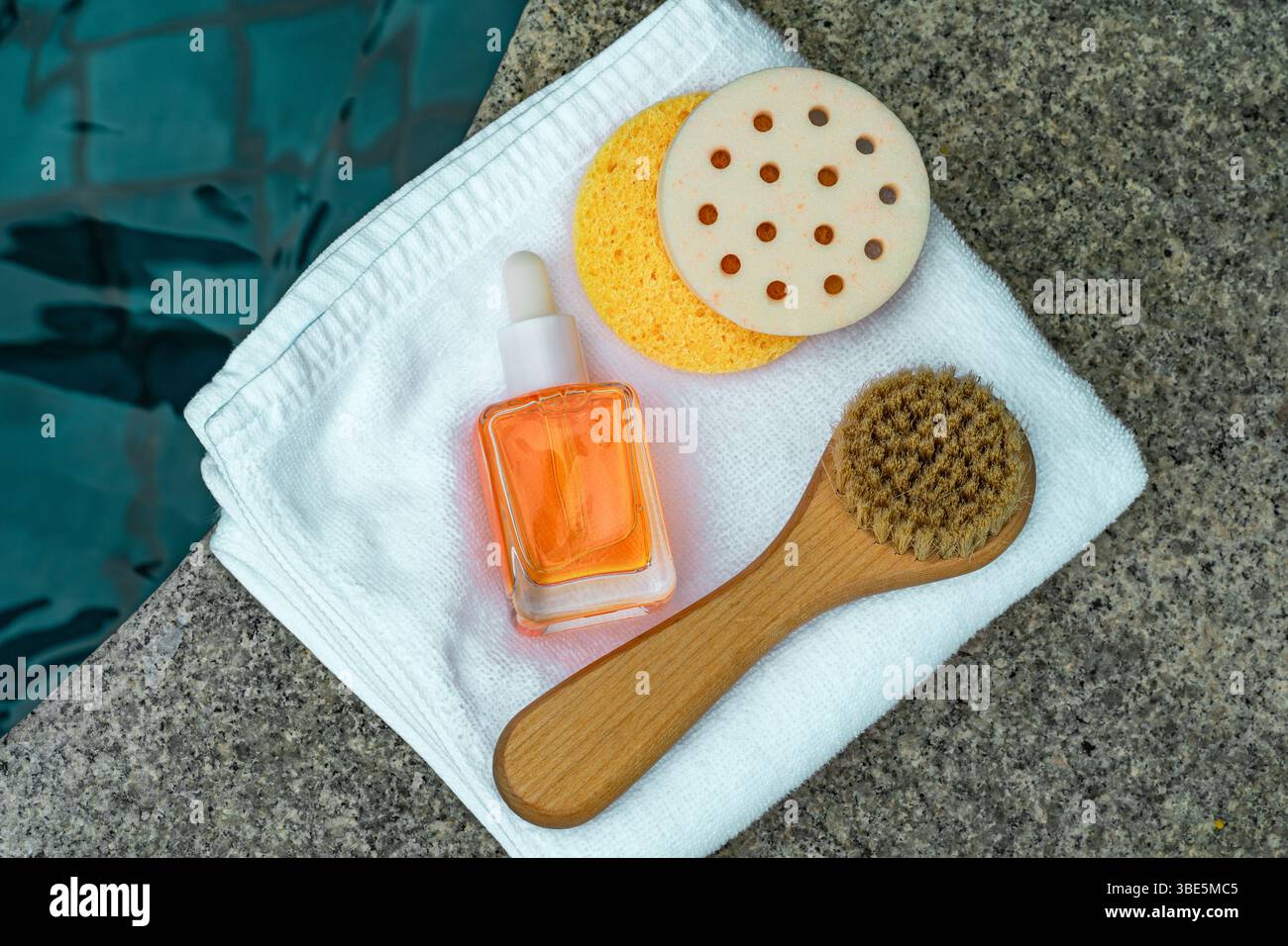 Blick von oben auf eine orangene Kosmetikflasche, Schwamm, Bürste und Handtuch, alle auf einer Steinoberfläche am türkisfarbenen Pool angeordnet. Stockfoto