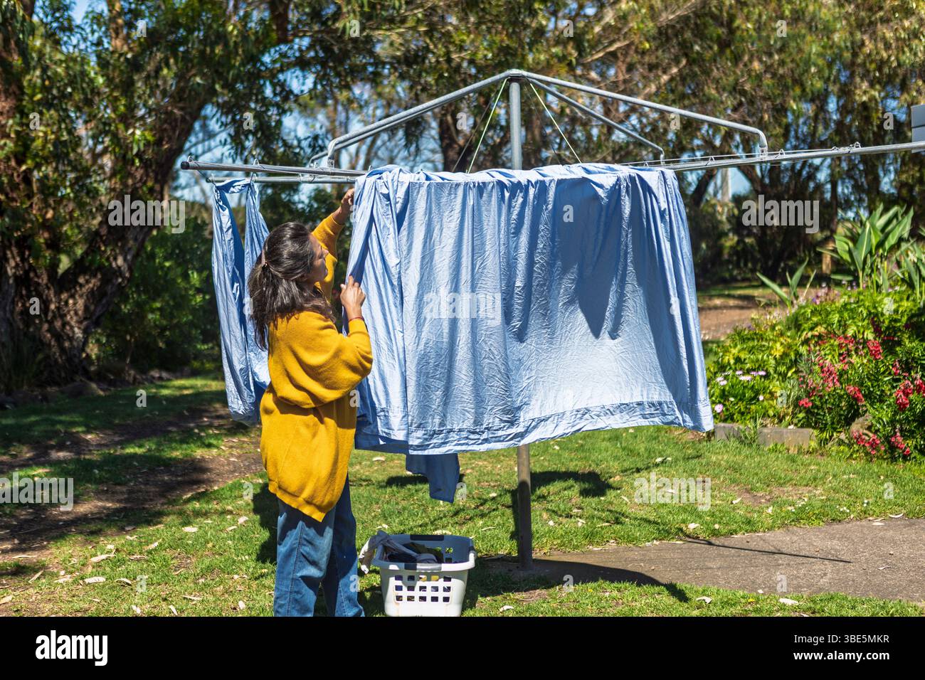 Eine Frau hängt Laken an einer Hügellinie in ihrem Garten, Australien. Sie erledigt ihre täglichen Aufgaben, umgeben von üppiger Vegetation und darunter Stockfoto