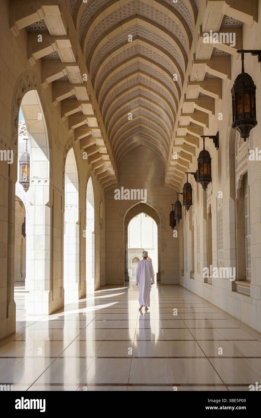 Rückansicht eines nicht erkennbaren Mannes in traditioneller Dschdasha-omanischer Kleidung, der entlang eines Korridors in der Sultan-Qaboos-Moschee in Maskat, Oman, spaziert Stockfoto