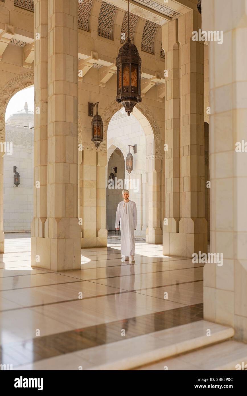 Ein Mann in traditioneller omanischer Dschasha-Kleidung spaziert durch den großen Korridor der Sultan-Qaboos-Moschee in Muscat, eingerahmt von kunstvollen Bögen und hängenden La Stockfoto