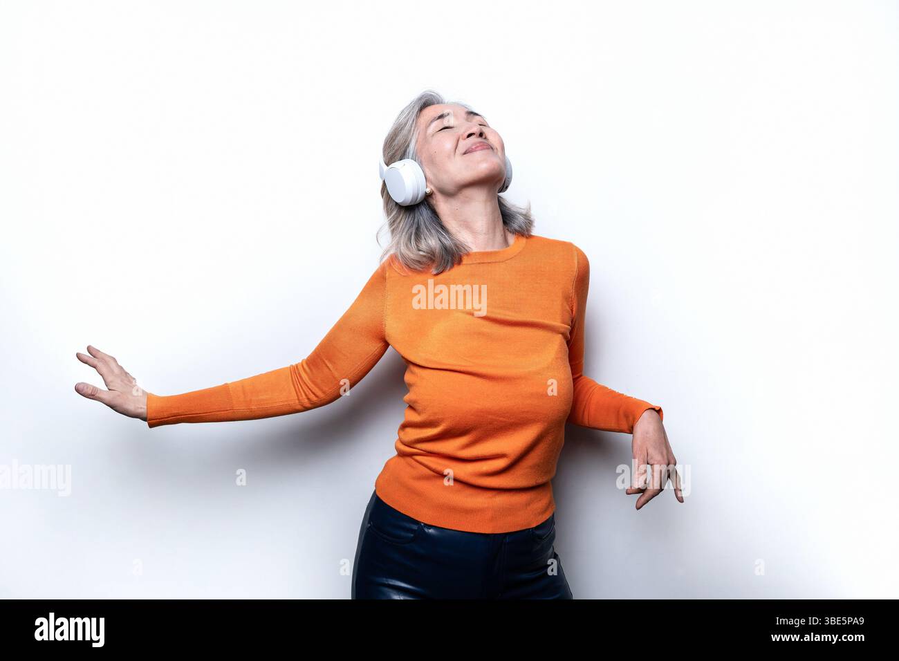 Asiatische Chinesische Seniorin mit Kopfhörern und einem orangefarbenen Pullover, die Musik mit einem fröhlichen Ausdruck genießt und vor einem schlichten weißen Hintergrund steht Stockfoto